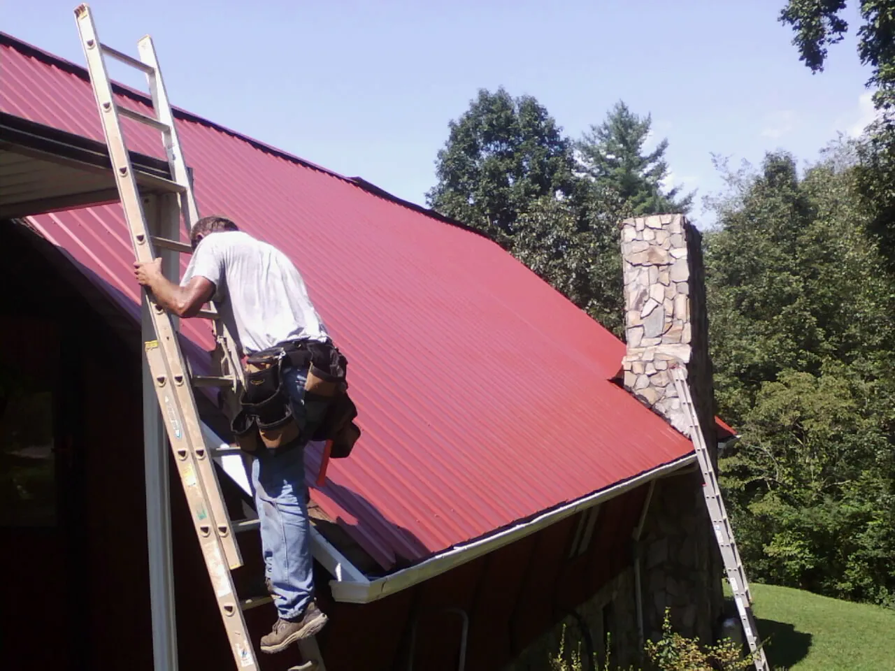 Roofer on a ladder beside a red metal roof and stone chimney — Soffit & Fascia Repair by Roofing Brothers in San Dimas